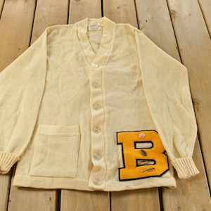 Pode incluir: Cardigã vintage creme com textura canelada, mangas compridas e decote em V. Possui um bolso de remendo e uma grande letra "B" amarela e preta na frente. Fechamento com botões.