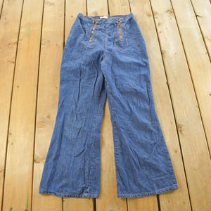 May include: Blue denim pants with a high waist and flared legs. The pants feature two decorative gold zippers on the front and a gold zipper closure. The pants are laid flat on a wooden surface.