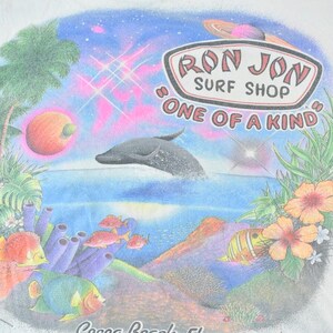 Puede incluir: Camiseta blanca con un gr&aacute;fico colorido que representa una escena de oc&eacute;ano tropical. El dise&ntilde;o incluye un delf&iacute;n, peces, coral y el texto "Ron Jon Surf Shop, One of a Kind, Cocoa Beach, FL."