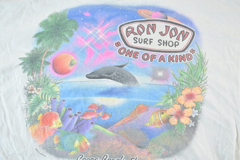 Puede incluir: Camiseta blanca con un gr&aacute;fico colorido que representa una escena de oc&eacute;ano tropical. El dise&ntilde;o incluye un delf&iacute;n, peces, coral y el texto "Ron Jon Surf Shop, One of a Kind, Cocoa Beach, FL."