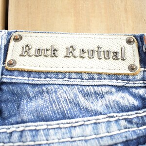 K&ouml;nnte beinhalten: Nahaufnahme einer blauen Jeanshose mit wei&szlig;en N&auml;hten. Die Jeans hat einen Leder-Patch mit dem Schriftzug "Rock Revival" in silberner Schrift.
