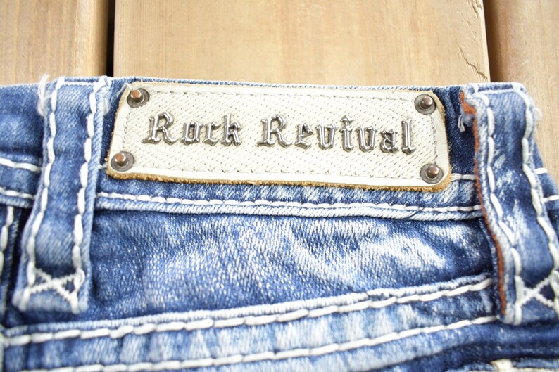 K&ouml;nnte beinhalten: Nahaufnahme einer blauen Jeanshose mit wei&szlig;en N&auml;hten. Die Jeans hat einen Leder-Patch mit dem Schriftzug "Rock Revival" in silberner Schrift.