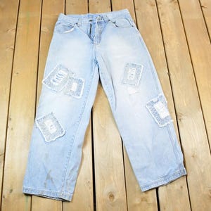 Peut inclure: Jean clair en denim avec des patchs déchirés. Le jean a une coupe décontractée et une conception classique à cinq poches. Les patchs sont rectangulaires et cousus sur le jean. Le jean est posé à plat sur une surface en bois.