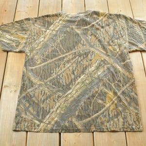 Puede incluir: Una camiseta de manga corta con un estampado de camuflaje en tonos marrones, verdes y beige. La camiseta est&aacute; hecha de un material suave y est&aacute; colocada sobre una superficie de madera. El dise&ntilde;o de camuflaje es detallado y cubre toda la prenda.
