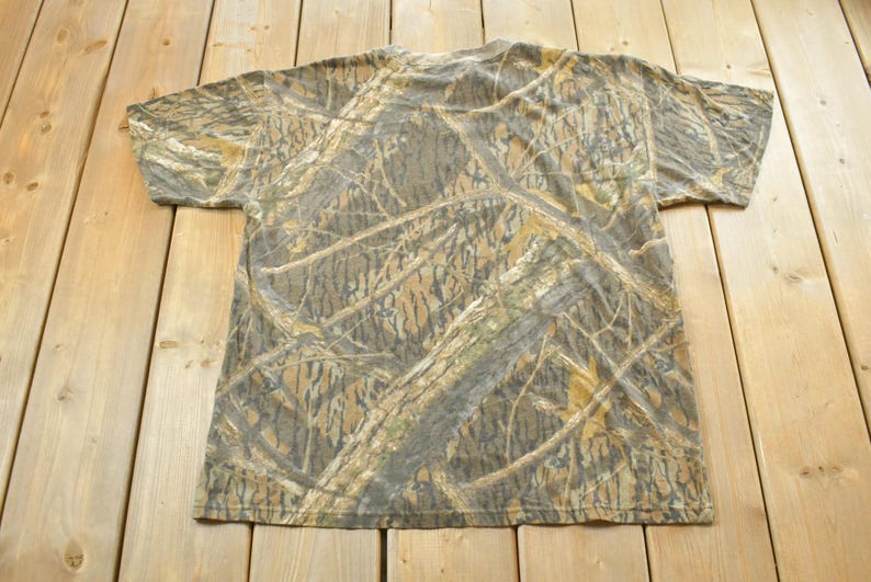 Puede incluir: Una camiseta de manga corta con un estampado de camuflaje en tonos marrones, verdes y beige. La camiseta est&aacute; hecha de un material suave y est&aacute; colocada sobre una superficie de madera. El dise&ntilde;o de camuflaje es detallado y cubre toda la prenda.