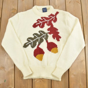 May include: A cream-colored knitted sweater with a brown, green, and red acorn design.