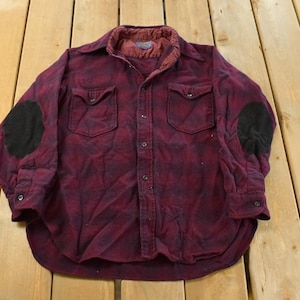 May include: A burgundy and black plaid button-down shirt with black elbow patches. The shirt has two chest pockets and a quilted lining. The shirt is laid flat on a wooden surface.