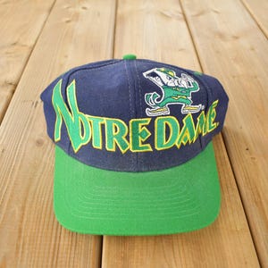 Può includere: Un cappello da baseball blu e verde con la parola "Notre Dame" ricamata in giallo e verde. Un folletto dei cartoni animati è ricamato sulla parte anteriore del cappello.