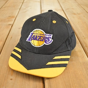 Peut inclure: Casquette de baseball noire avec le logo des Lakers de Los Angeles en violet, jaune et blanc. La casquette a une visière jaune et des rayures jaunes sur le côté. Fabriquée en tissu résistant.