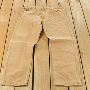 Könnte beinhalten: Beigefarbene Arbeitshose mit geradem Bein. Die Hose besteht aus strapazierfähigem Material und verfügt über mehrere Taschen sowie einen Knopf- und Reißverschluss. Die Hose liegt flach auf einer Holzoberfläche.