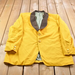 Könnte beinhalten: Ein gelber Blazer mit einem braunen Samtkragen. Der Blazer hat goldene Knöpfe und liegt flach auf einer Holzoberfläche.