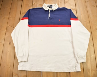 May include: A white long-sleeve rugby shirt with a blue and red stripe across the chest. The shirt has a button-up collar and a small embroidered logo on the left chest.