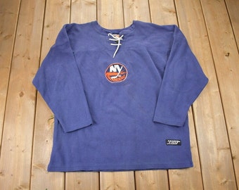 May include: A blue long-sleeved sweatshirt with a white lace-up neckline. The sweatshirt features an orange and white New York Islanders logo with the text "NY Islanders". The sweatshirt also has a black tag with the text "Zit" on the bottom right side.