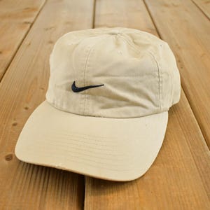 Op de afbeelding: Een beige baseballpet met een gebogen klep. De pet heeft een zwart Nike swoosh-logo. De hoed is gemaakt van een zachte, licht versleten stof en staat op een houten achtergrond.