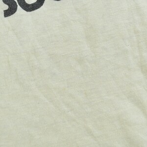 May include: A light cream-colored fabric garment featuring a portion of black text printed at the top. The visible text includes the letters H and S, which appear to be part of a larger word or phrase. The material has a visible woven texture.
