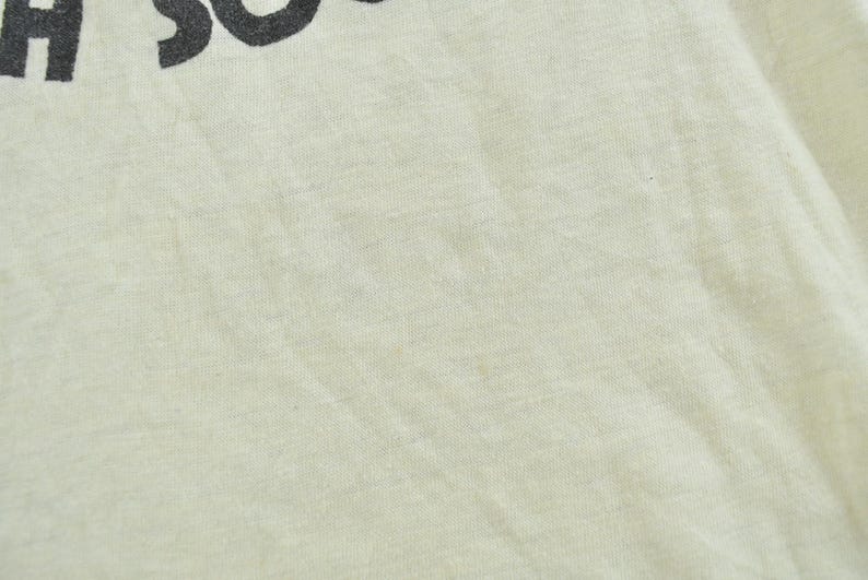 May include: A light cream-colored fabric garment featuring a portion of black text printed at the top. The visible text includes the letters H and S, which appear to be part of a larger word or phrase. The material has a visible woven texture.