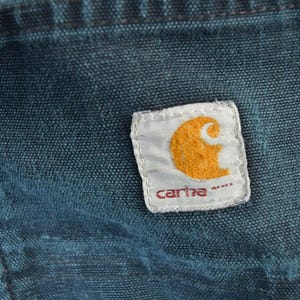 May include: Close-up of a section of blue denim fabric with a Carhartt brand patch. The patch is white with an orange logo and the brand name in red. The denim shows signs of wear.