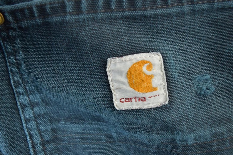 May include: Close-up of a section of blue denim fabric with a Carhartt brand patch. The patch is white with an orange logo and the brand name in red. The denim shows signs of wear.