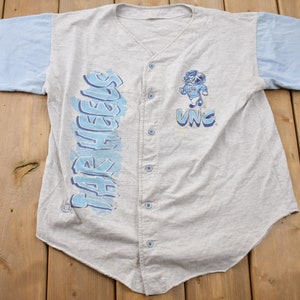 May include: A gray and light blue baseball jersey with the words "Tar Heels" printed on the left side and "UNC" printed on the right side. A cartoon mascot is printed on the right side of the jersey.