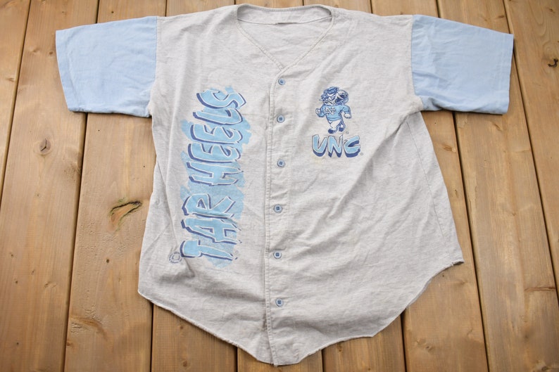 May include: A gray and light blue baseball jersey with the words "Tar Heels" printed on the left side and "UNC" printed on the right side. A cartoon mascot is printed on the right side of the jersey.