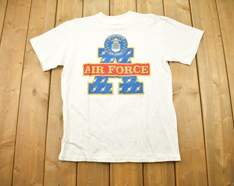 May include: White t-shirt with a blue and gold "Air Force" graphic on the back. The graphic features a stylised "A" with the Air Force emblem and the words "Air Force" in red.