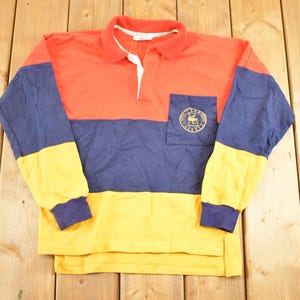 Peut inclure: Une chemise à manches longues avec blocs de couleurs rouge, bleu marine et jaune. La chemise a un col blanc, une petite poche avec un emblème doré et des poignets bleu marine. La chemise est en matière souple.