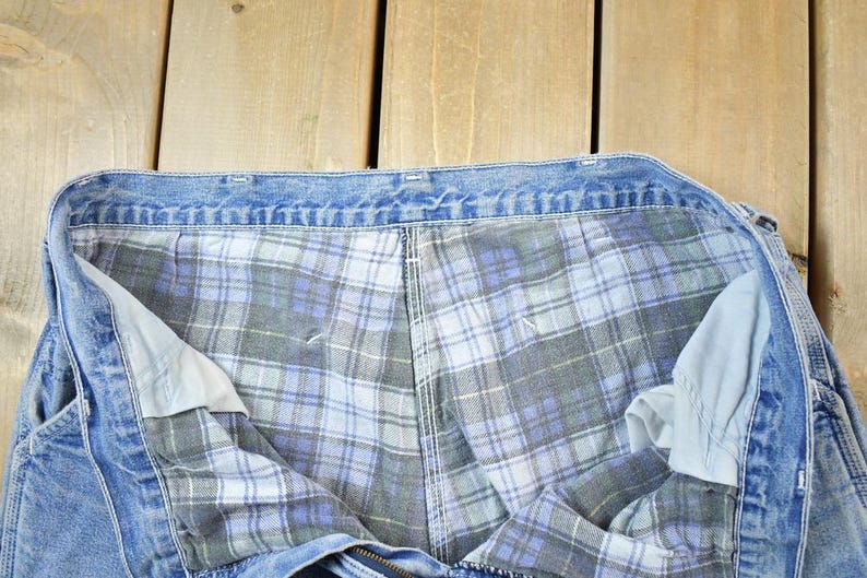 K&ouml;nnte beinhalten: Nahaufnahme einer blauen Jeans-Arbeitshose mit einem karierten Flanellfutter in Blau-, Grau- und Wei&szlig;t&ouml;nen. Die Hose liegt flach, wobei das Innenfutter und das Jeans-Au&szlig;enmaterial zu sehen sind.