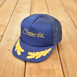 Op de afbeelding: Een marineblauwe truckerpet met een mesh achterkant. De pet heeft het woord "Timberlea" in gouden letters over de voorkant. Gouden bladmotieven sieren de rand. De pet staat tegen een houten achtergrond.