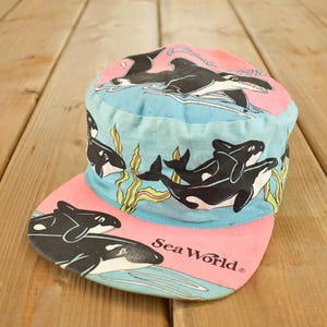 Può includere: Un cappellino da baseball vintage di Sea World con visiera rosa e corona azzurra. Il cappellino è decorato con orche bianche e nere e il logo Sea World.