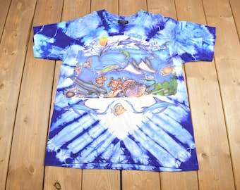 Peut inclure: Un t-shirt bleu tie-dye avec un imprimé coloré de différentes créatures marines, notamment des dauphins, des poissons et une tortue. L'imprimé est centré sur le t-shirt et entouré d'un motif tie-dye bleu et blanc.