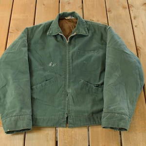 May include: A vintage green work jacket with a full zip, two front pockets, and a collared neckline. The jacket shows signs of wear, with some discoloration and markings. The interior lining is a contrasting brown colour.