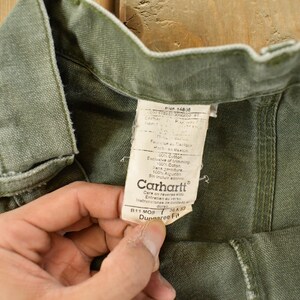 Puede incluir: Primer plano de pantalones Carhartt verde oliva con una etiqueta visible. La etiqueta muestra el logotipo de Carhartt e informaci&oacute;n del producto. Los pantalones est&aacute;n hechos de una tela duradera y las costuras son visibles. Los pantalones son sostenidos por una persona.