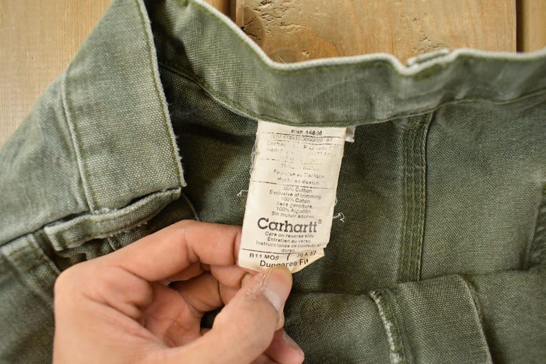Puede incluir: Primer plano de pantalones Carhartt verde oliva con una etiqueta visible. La etiqueta muestra el logotipo de Carhartt e informaci&oacute;n del producto. Los pantalones est&aacute;n hechos de una tela duradera y las costuras son visibles. Los pantalones son sostenidos por una persona.