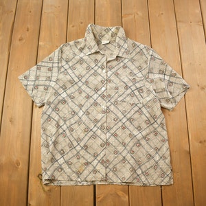 May include: A beige short-sleeved button-down shirt with a geometric pattern in shades of brown, black, and red.