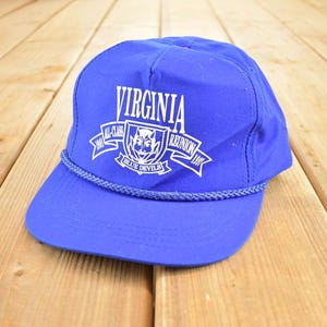 Puede incluir: Gorra de béisbol azul con texto blanco "VIRGINIA" y "ALL-CLASS REUNION 1997 BLUE DEVILS". La gorra tiene un detalle de cuerda alrededor del borde y está sobre un fondo de madera.