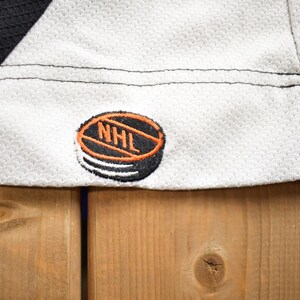 Puede incluir: Jersey de hockey blanco con ribetes negros y dorados. El jersey tiene un parche de logotipo de la NHL negro y naranja en el lado inferior derecho.
