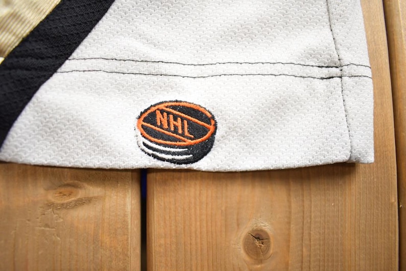 Puede incluir: Jersey de hockey blanco con ribetes negros y dorados. El jersey tiene un parche de logotipo de la NHL negro y naranja en el lado inferior derecho.