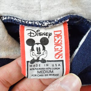 K&ouml;nnte beinhalten: Nahaufnahme eines Disney Designs-Etiketts auf einem marineblauen Kapuzenpullover. Das rot-wei&szlig;e Etikett zeigt Mickey Mouse. Das Etikett lautet "MADE IN U.S.A." und "MEDIUM".