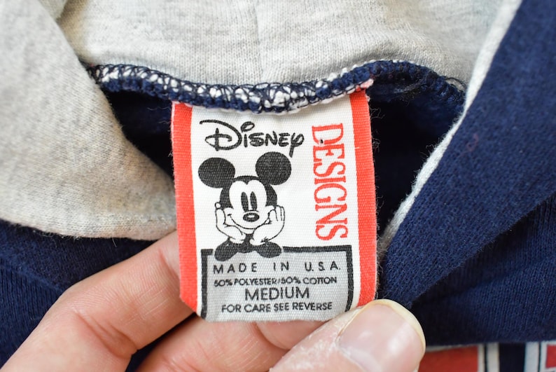 K&ouml;nnte beinhalten: Nahaufnahme eines Disney Designs-Etiketts auf einem marineblauen Kapuzenpullover. Das rot-wei&szlig;e Etikett zeigt Mickey Mouse. Das Etikett lautet "MADE IN U.S.A." und "MEDIUM".