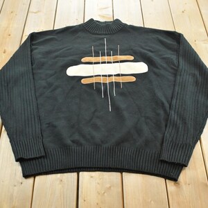Könnte beinhalten: Ein schwarzer Langarmpullover mit Stehkragen sowie gerippten Bündchen an Ärmeln und Saum. Die Vorderseite zeigt ein zentrales grafisches Design aus horizontalen, beigen und cremefarbenen Applikationen mit feinen, vertikalen weißen Stickereien.