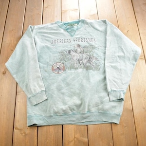 May include: A light blue sweatshirt with a graphic of three dogs running through a field. The text "American Sportsmen" is printed above the graphic. The graphic also includes a small circle with the text "Great American Outdoors & Tourism".