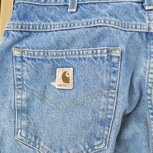 Puede incluir: Primer plano de la parte trasera de unos vaqueros de mezclilla azul claro. Los vaqueros tienen un bolsillo trasero con un parche con el logotipo de Carhartt marr&oacute;n y beige. Las costuras son visibles en hilo amarillo y blanco.