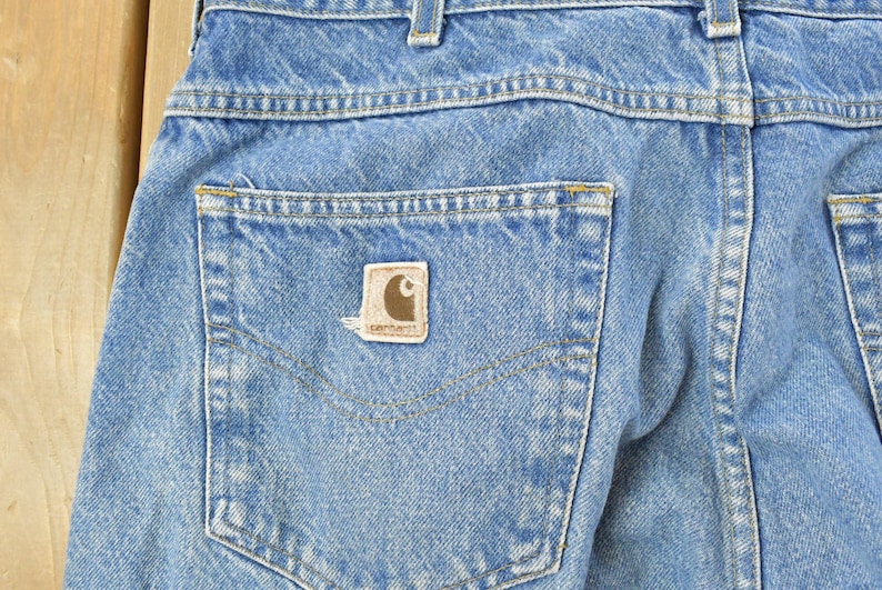 Puede incluir: Primer plano de la parte trasera de unos vaqueros de mezclilla azul claro. Los vaqueros tienen un bolsillo trasero con un parche con el logotipo de Carhartt marr&oacute;n y beige. Las costuras son visibles en hilo amarillo y blanco.