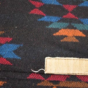 May include: Close-up of a black textile with a geometric pattern in red, blue, orange, and green. A rectangular beige tag is sewn onto the fabric. The textile has a woven texture.