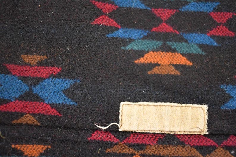 May include: Close-up of a black textile with a geometric pattern in red, blue, orange, and green. A rectangular beige tag is sewn onto the fabric. The textile has a woven texture.