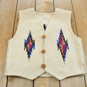 May include: A sleeveless vest in an off-white color featuring a V-neckline and three brown circular buttons down the center. The front panels are decorated with two symmetrical diamond-shaped patterns in blue, red, and black thread.