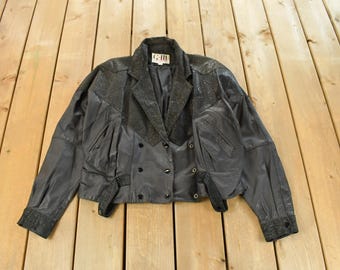 May include: A vintage black leather jacket with a double-breasted design. The jacket features a notched collar, long sleeves, and decorative accents. The jacket is displayed on a wooden surface.