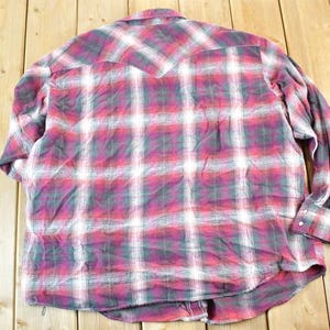 Puede incluir: Una camisa de cuadros de manga larga en tonos rojos, blancos y grises. La camisa tiene una parte delantera abotonada, un cuello y pu&ntilde;os abotonados. La parte trasera de la camisa presenta un detalle de canes&uacute;. La tela parece ser una franela suave.