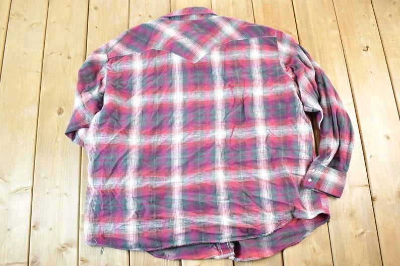 Puede incluir: Una camisa de cuadros de manga larga en tonos rojos, blancos y grises. La camisa tiene una parte delantera abotonada, un cuello y pu&ntilde;os abotonados. La parte trasera de la camisa presenta un detalle de canes&uacute;. La tela parece ser una franela suave.