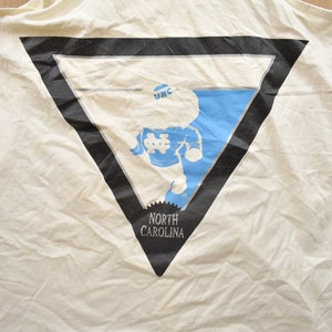 May include: White tank top with a black triangle graphic featuring a cartoon mascot wearing a blue and white jersey with the text "UNC" on the hat and "NORTH CAROLINA" below the graphic.